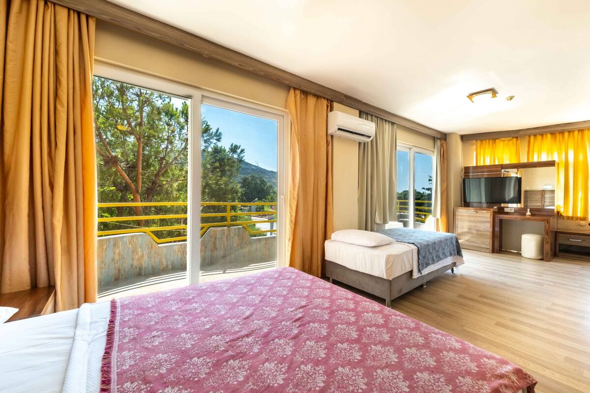 Hotelkamer met een bed, een bureau, een televisie en een balkon met uitzicht op bomen en een berg.