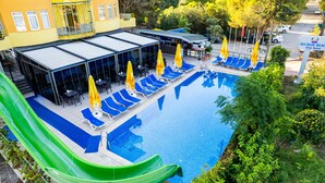 Une piscine avec des chaises longues bleues et des parasols jaunes, un toboggan vert et un espace abrité avec des tables et des chaises.