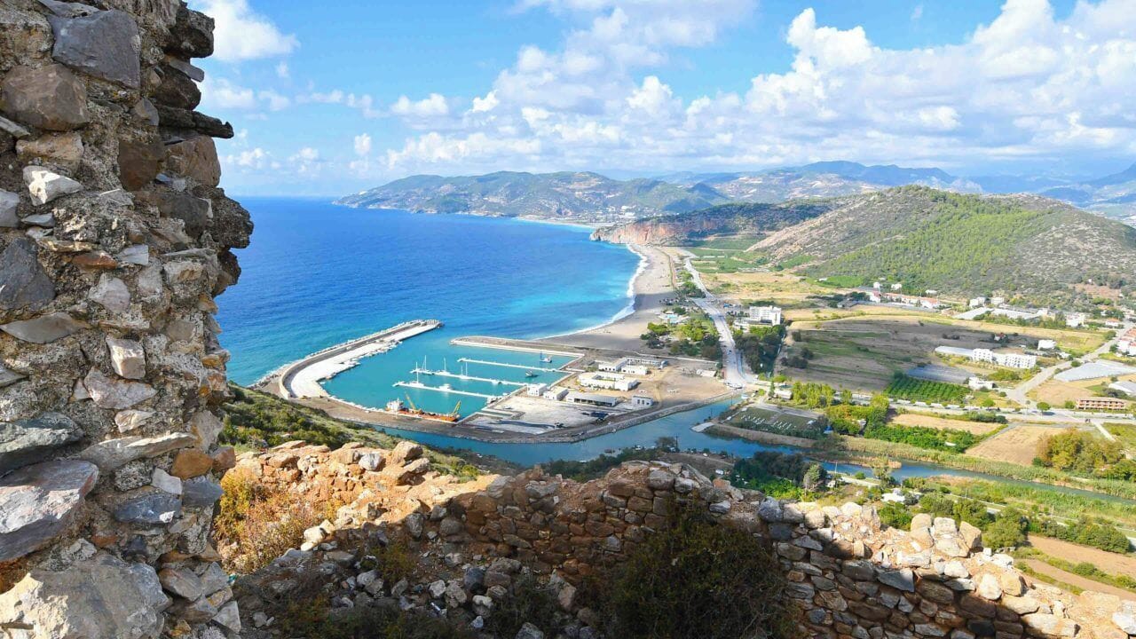 Blick auf die Küste mit einer Steinmauer, einem Yachthafen und einer hügeligen Landschaft.