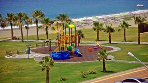 Un terrain de jeux avec un toboggan et des structures d’escalade, entouré de palmiers et de bancs, avec une plage et des parasols en arrière-plan.