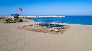 Une plage avec des chaises longues et des parasols sous un abri, une tour de surveillance pour les sauveteurs et une jetée surmontée d’un phare.