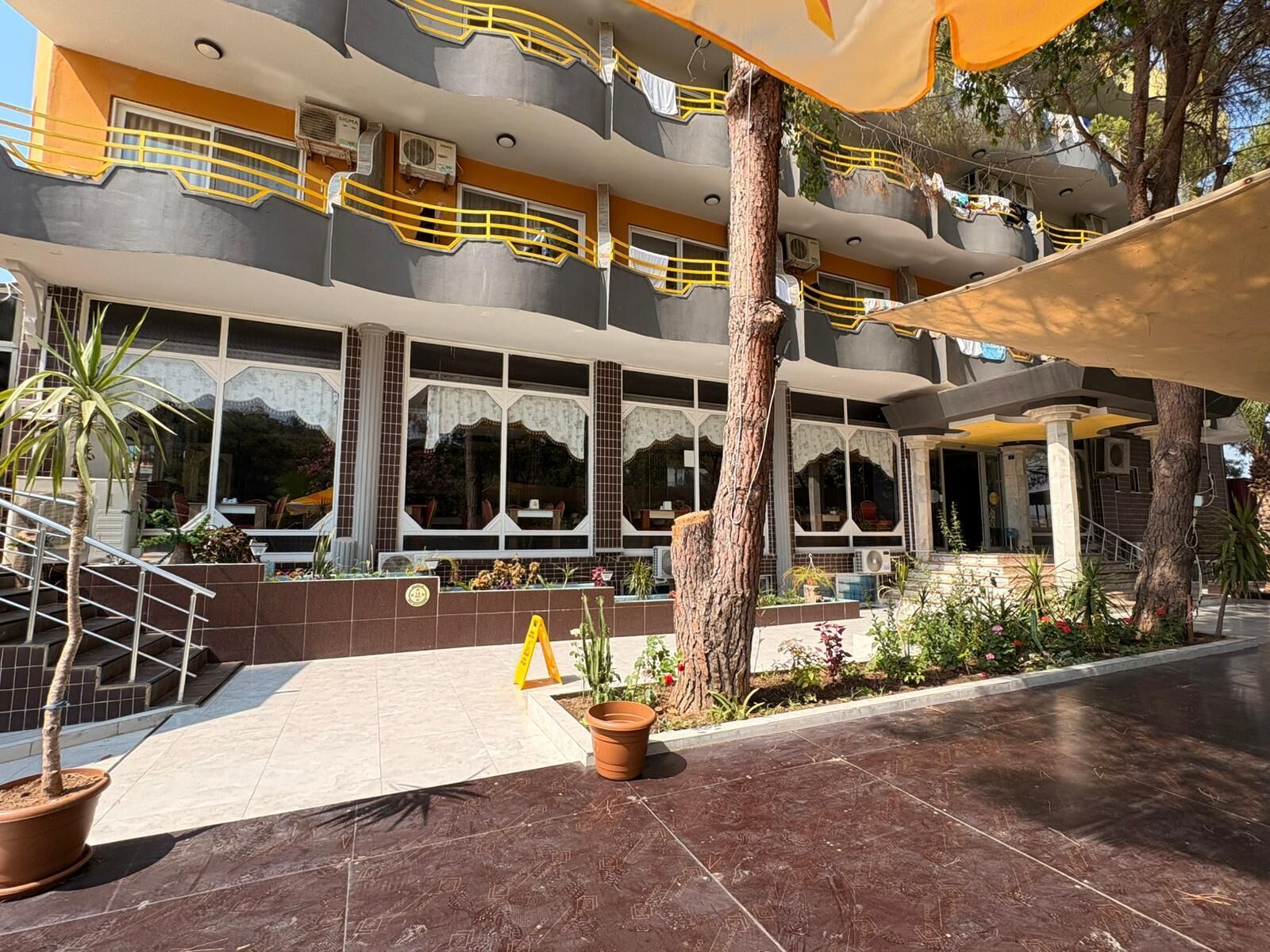 Un hotel de varios pisos con un diseño moderno, que cuenta con balcones con barandillas amarillas, ventanas grandes y un área exterior bien cuidada con plantas en macetas y un patio con baldosas.