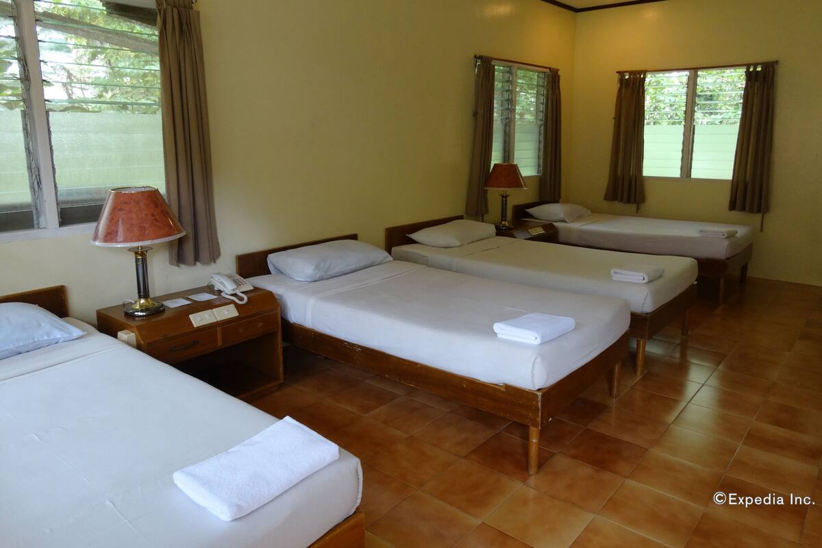 Quarto de hotel com quatro camas de solteiro, uma mesa de cabeceira de madeira com abajur, telefone e janela com cortinas.