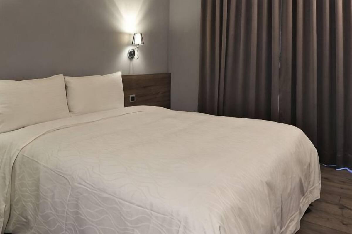A neatly made bed with a headboard, a wall-mounted light fixture, and a window with curtains.