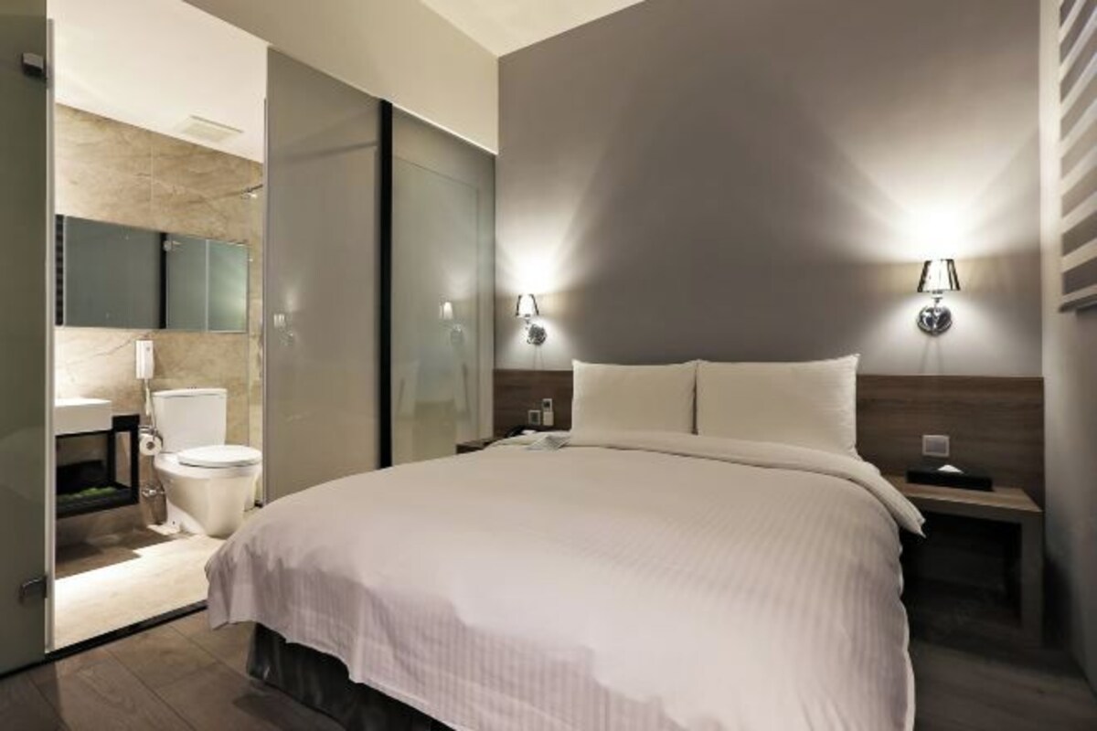 A modern hotel room with a large bed, a bedside table, a nightstand, a wall-mounted lamp, and a bathroom with a toilet and sink.