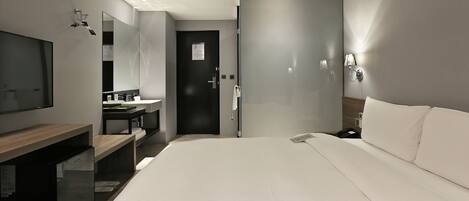 A modern hotel room with a large bed, a desk with a flat-screen TV, a mirror, a bathroom with a shower, and a door to another room.