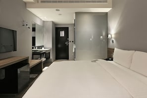A modern hotel room with a large bed, a desk with a flat-screen TV, a mirror, a bathroom with a shower, and a door to another room.