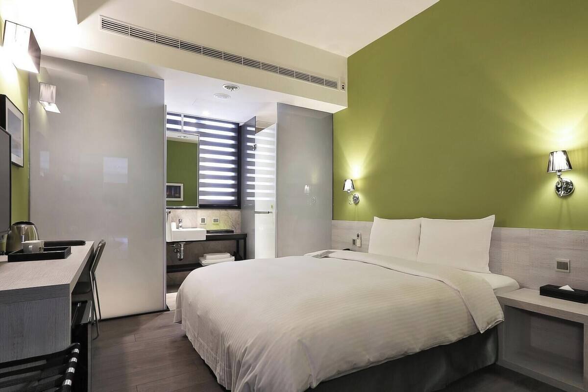 A hotel room with a bed, bedside table, desk, and a bathroom with a sink and mirror.