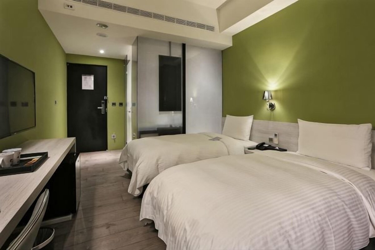 Habitación de hotel con dos camas, un escritorio, una silla, un espejo y una lámpara de pared.