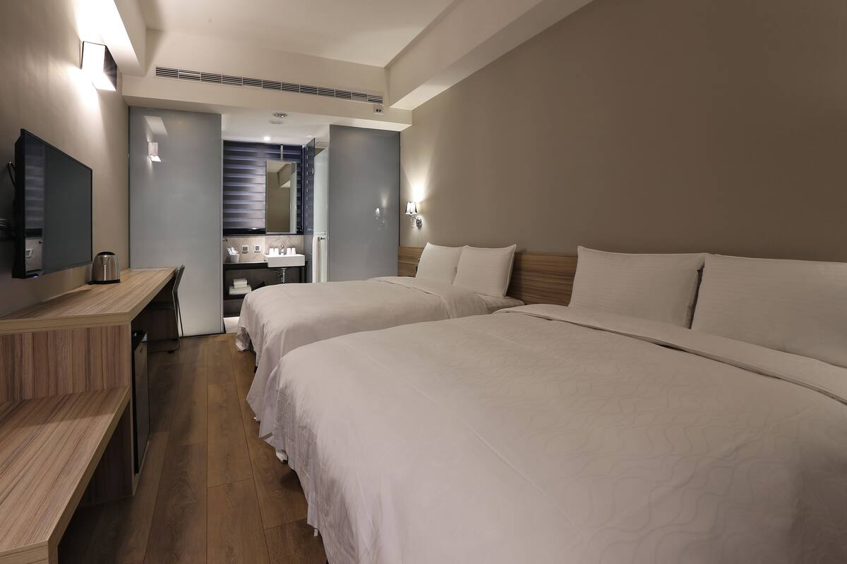 A hotel room with two beds, a desk, a TV, and a bathroom visible through an open door.