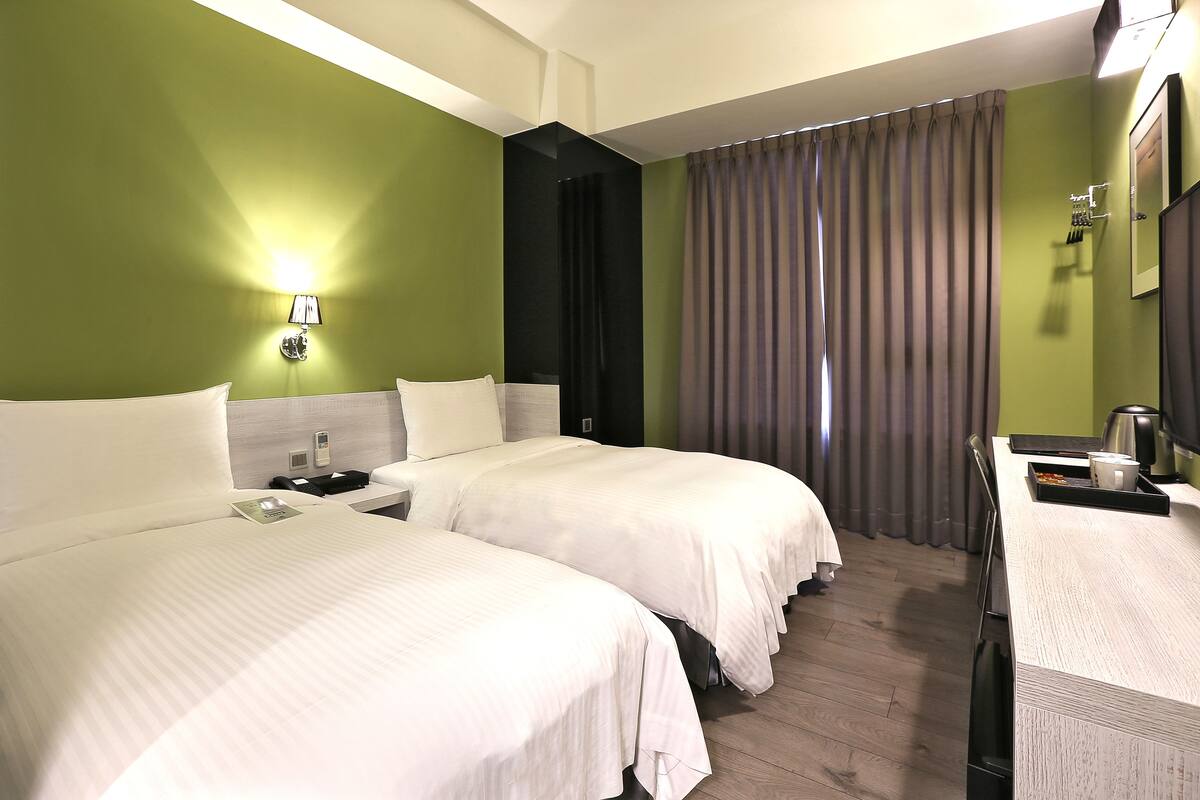 A hotel room with two beds, a desk, a mirror, a lamp, and a window with curtains.