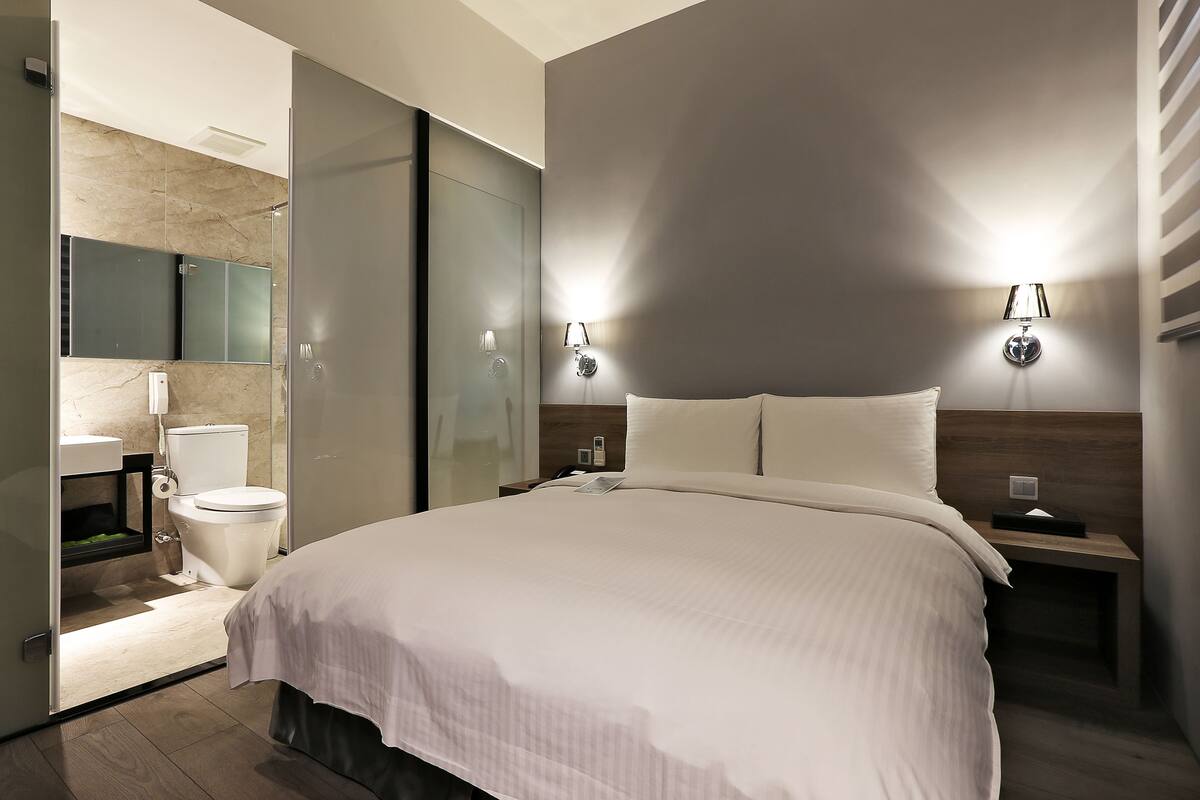 A modern hotel room with a large bed, a bedside table with a lamp, a nightstand, a mirror, a toilet, and a shower area.