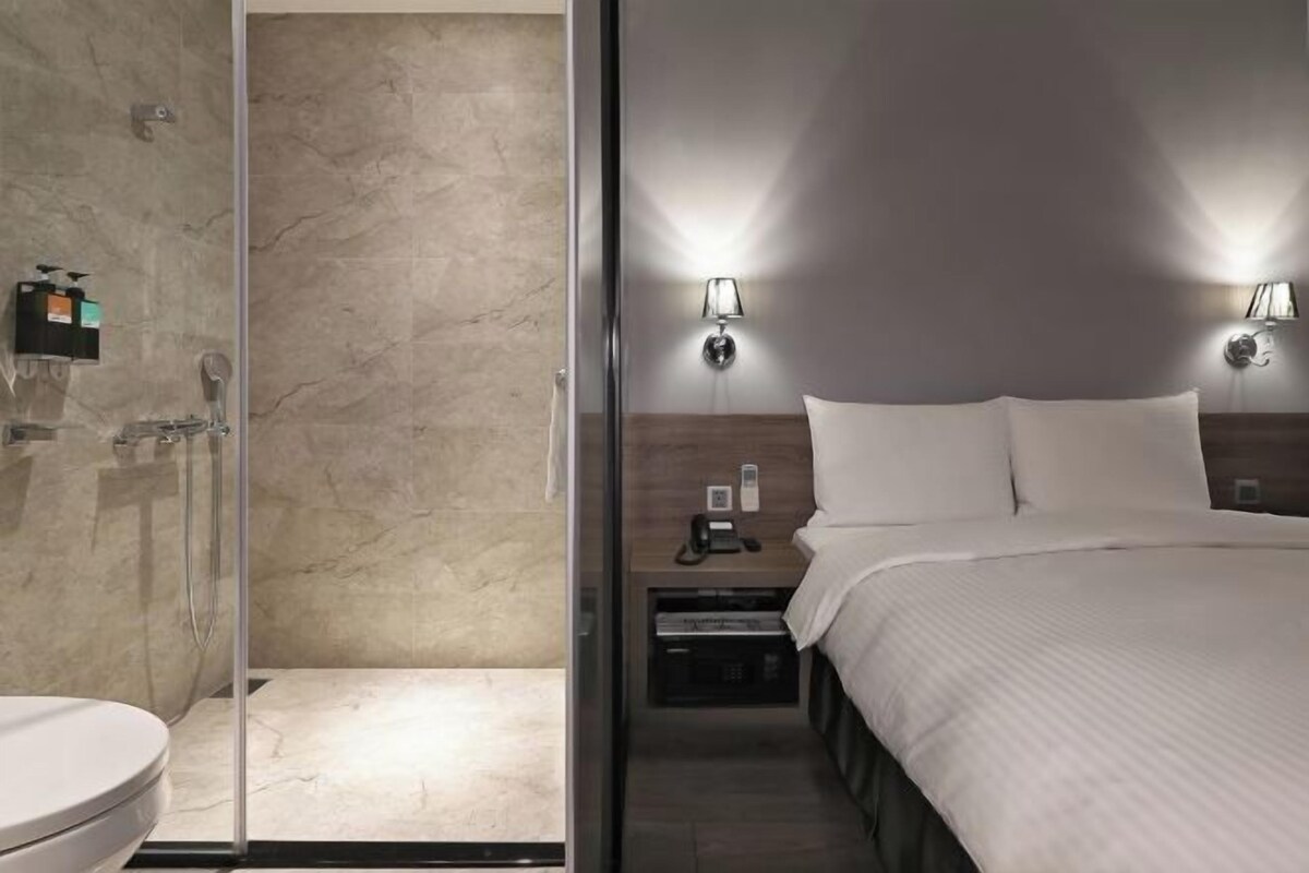 Una habitación de hotel moderna con cabina de ducha de vidrio, una mesita de noche con lámpara y una cama bien tendida.