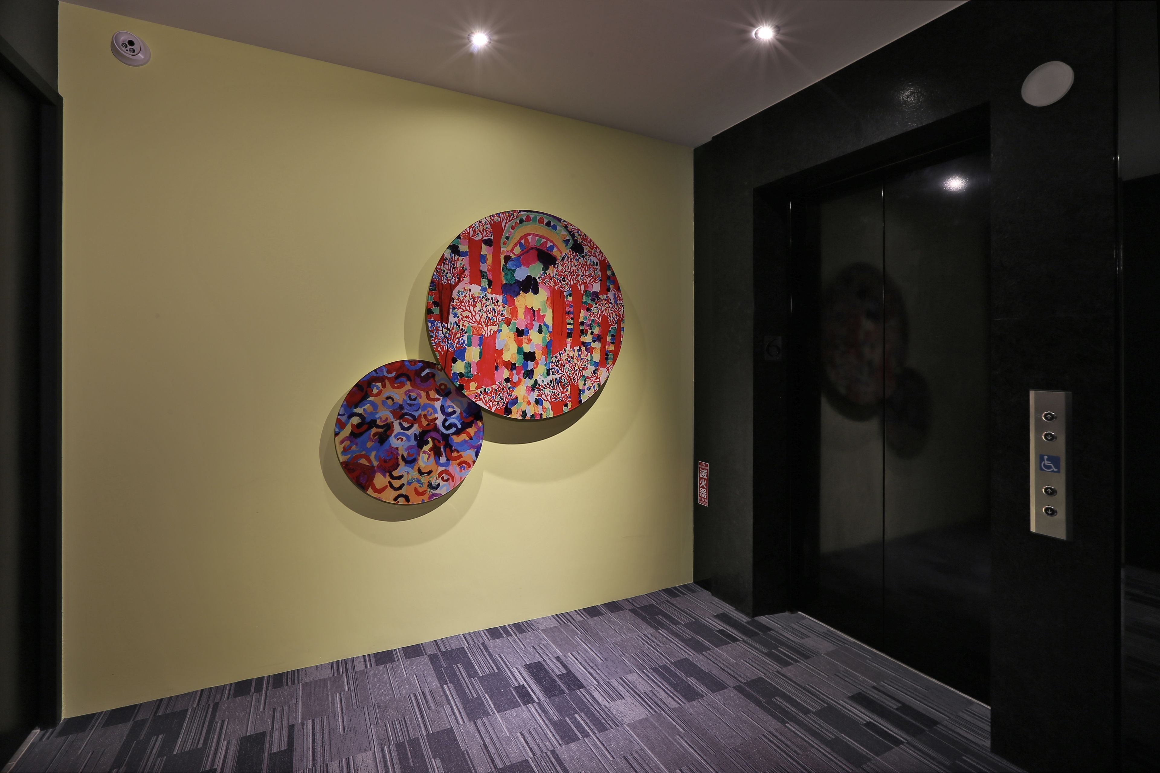 A hallway with a yellow wall and a large circular artwork. There is an elevator with a glass door and a control panel with buttons.