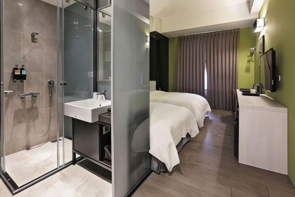 Habitación de hotel moderna con ducha de vidrio, baño separado con lavamanos, cama con ropa blanca y televisor de pantalla plana montado en la pared.