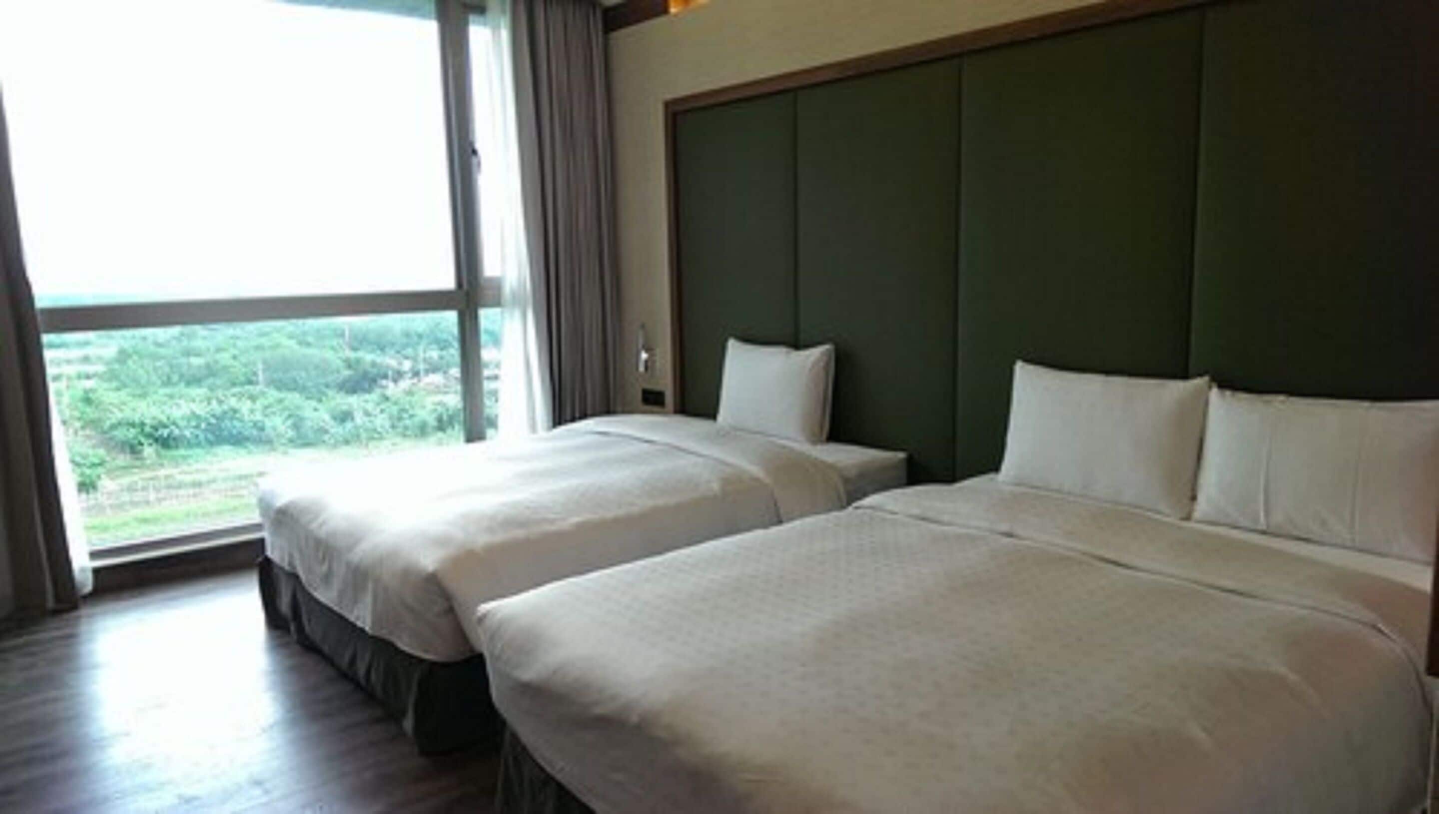 A hotel room with two beds, a large window, and a view of greenery.