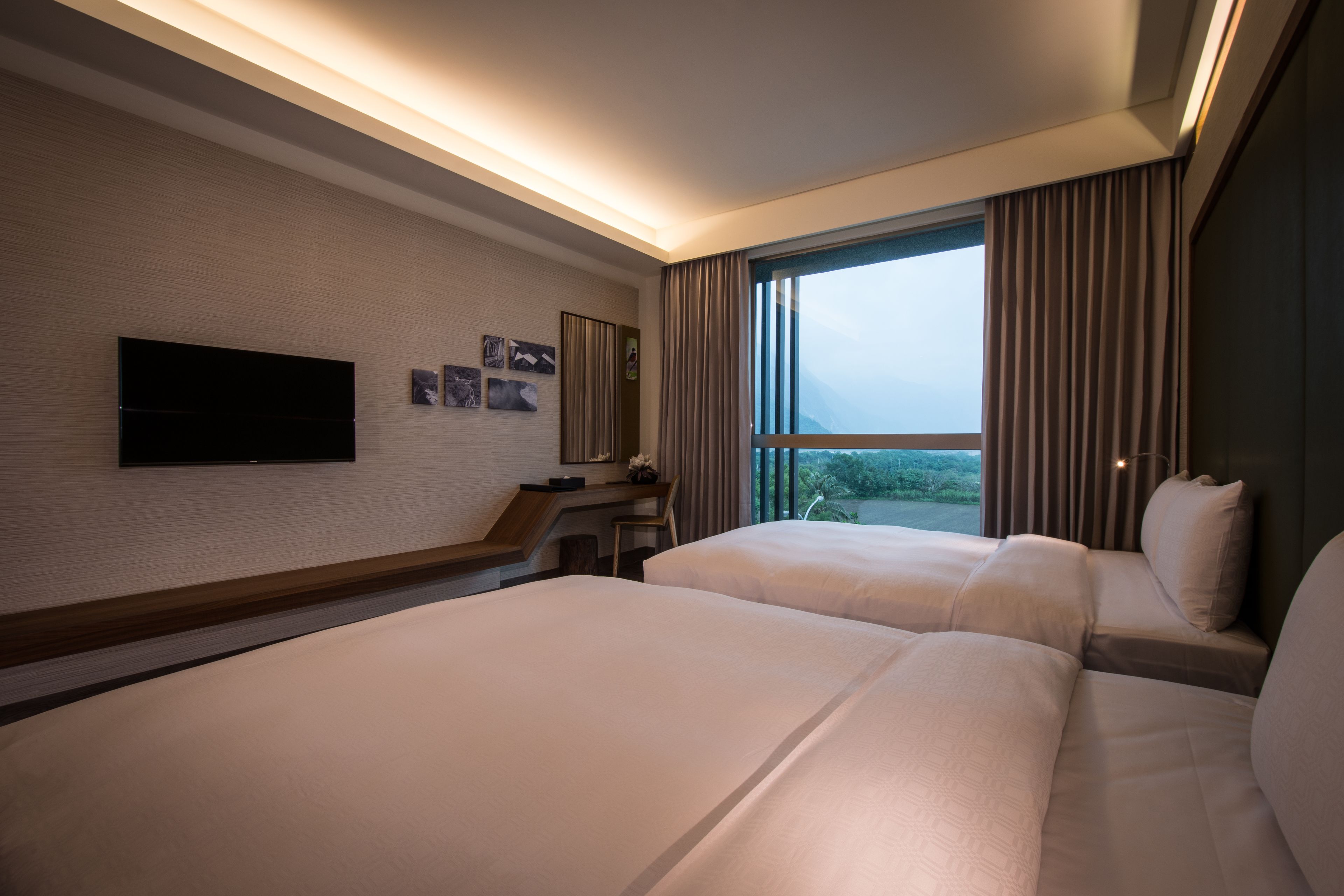 A hotel room with two beds, a flat-screen TV mounted on the wall, a desk, and a large window with curtains.