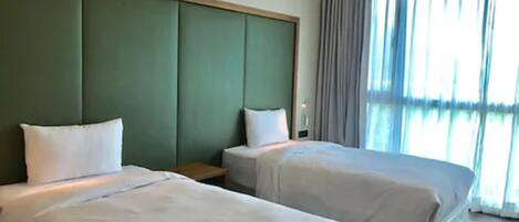 A hotel room with two beds, a green padded headboard, a bedside table, and a large window with curtains.