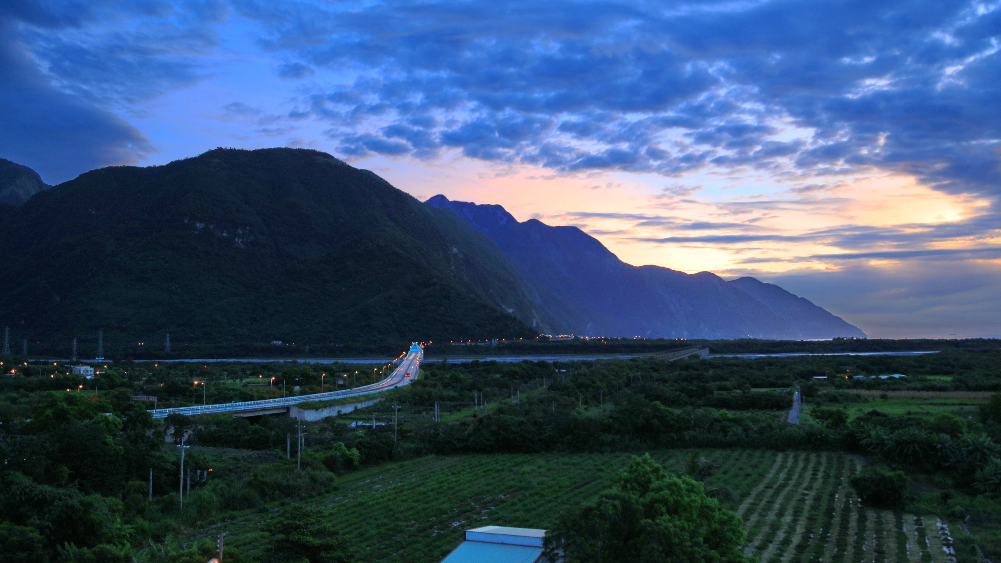 A mountainous landscape with a winding road, greenery, and a distant view of a mountain range.