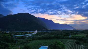 A mountainous landscape with a winding road, greenery, and a distant view of a mountain range.