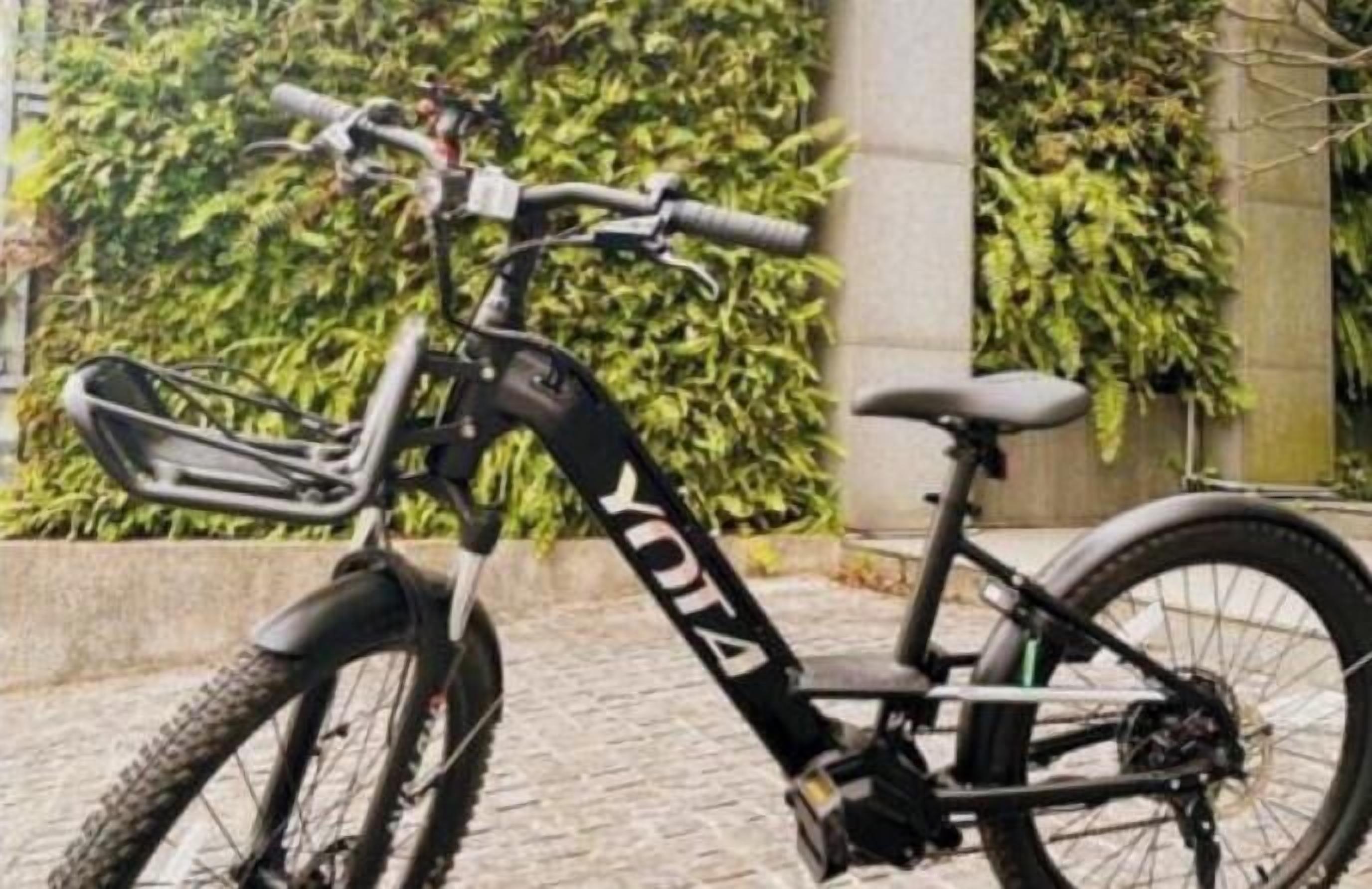 A stationary exercise bike with a black frame and "YOGA" branding, positioned outdoors with a stone wall and greenery in the background.