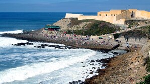 A coastal area with rocky terrain, a beach with people, and a large building with a lighthouse. - Hotel Neptuno (Peniche)