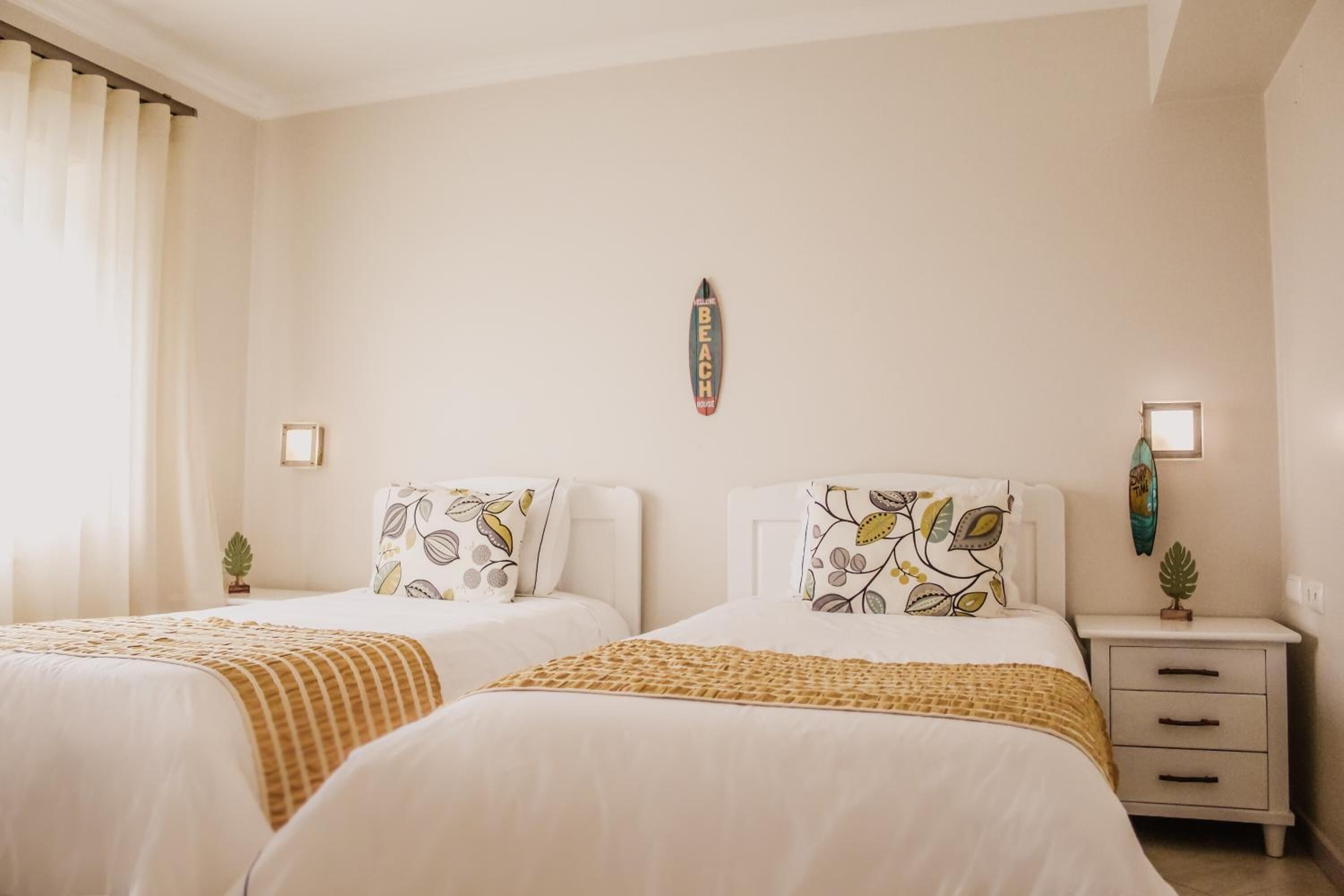 A hotel room with two beds, each with a patterned pillow and a beige throw blanket. There is a small bedside table with a plant and a framed picture.
