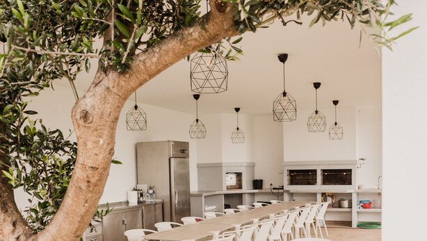 A spacious dining area with white tables and chairs, a large tree, and hanging pendant lights. - Hotel Neptuno (Peniche)