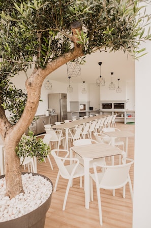 A spacious dining area with white tables and chairs, a large tree, and hanging pendant lights. - Hotel Neptuno (Peniche)