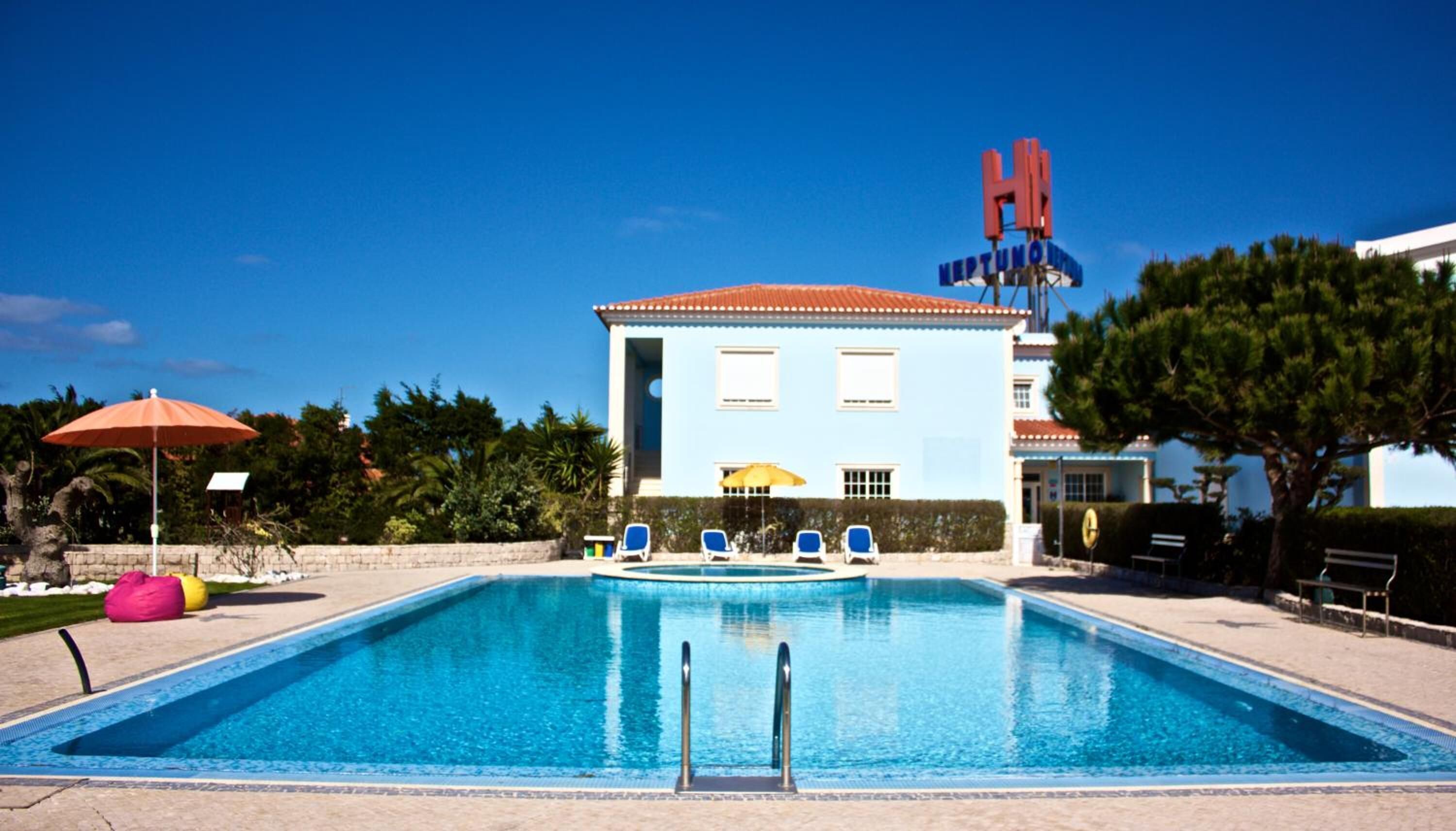 Uma piscina com espreguiçadeiras e guarda-sóis, um edifício com fachada azul e telhado vermelho, e uma placa com 'H' e 'NEPTUNIA' visíveis.