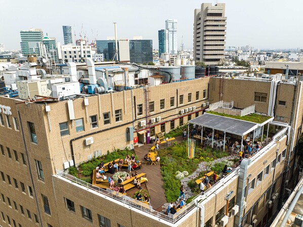 A rooftop garden with seating areas and a covered patio, surrounded by a cityscape with modern buildings.