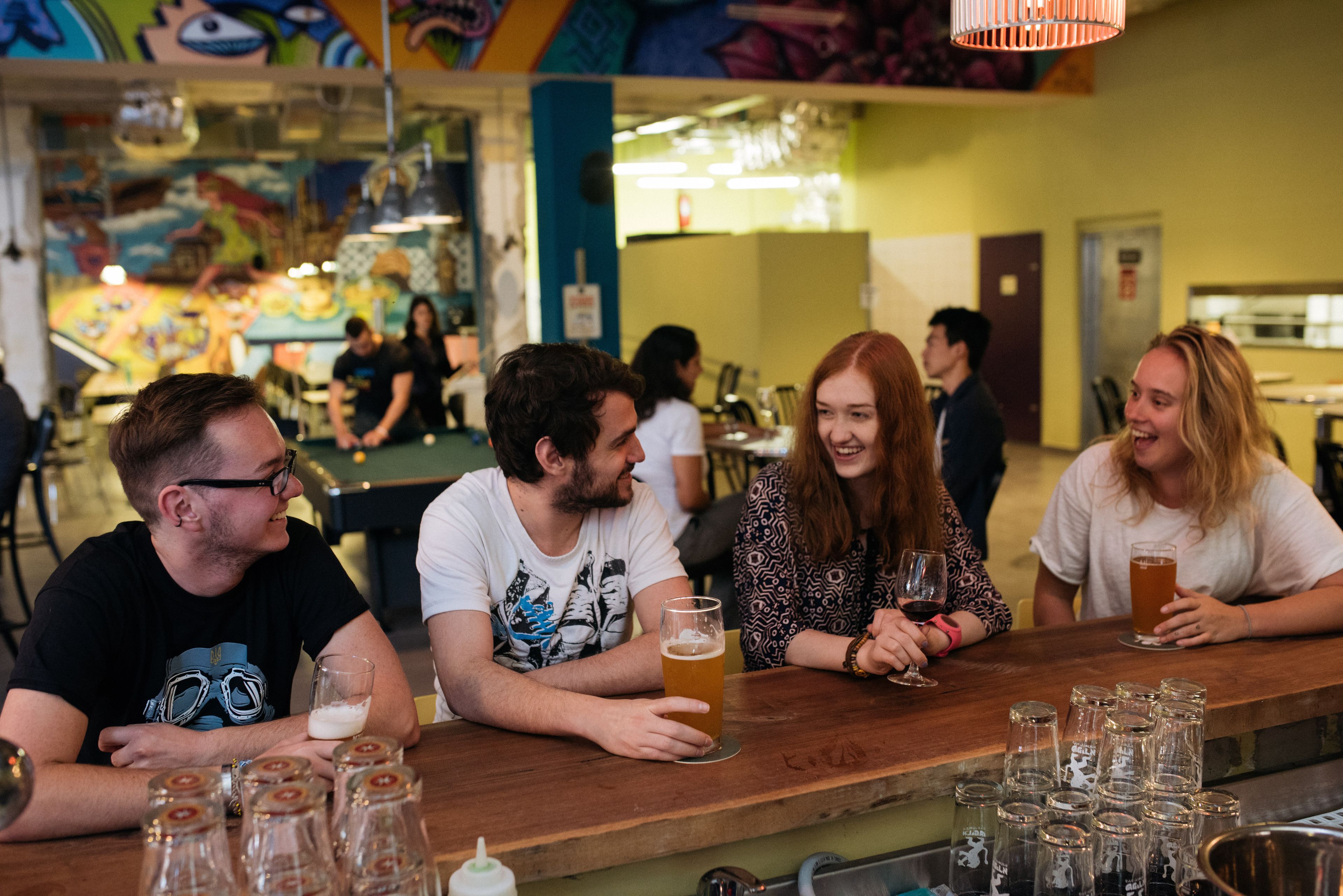 A bar area with people seated at tables, glasses of beer, and a variety of bottles on the counter.