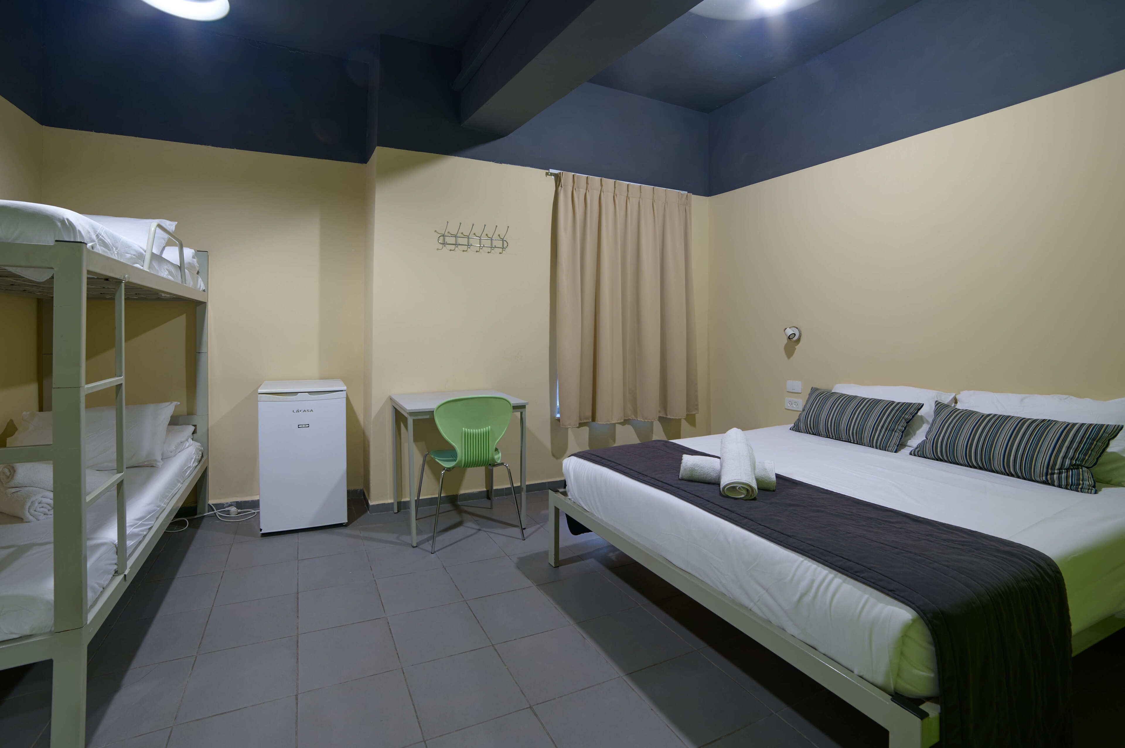 A dorm-style room with bunk beds, a desk, a chair, a small refrigerator, and a bed with pillows and a blanket.