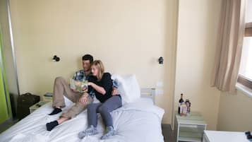 Une chambre d’hôtel avec un lit, une table de chevet sur laquelle se trouvent une bouteille et des verres, et une petite table avec des livres.