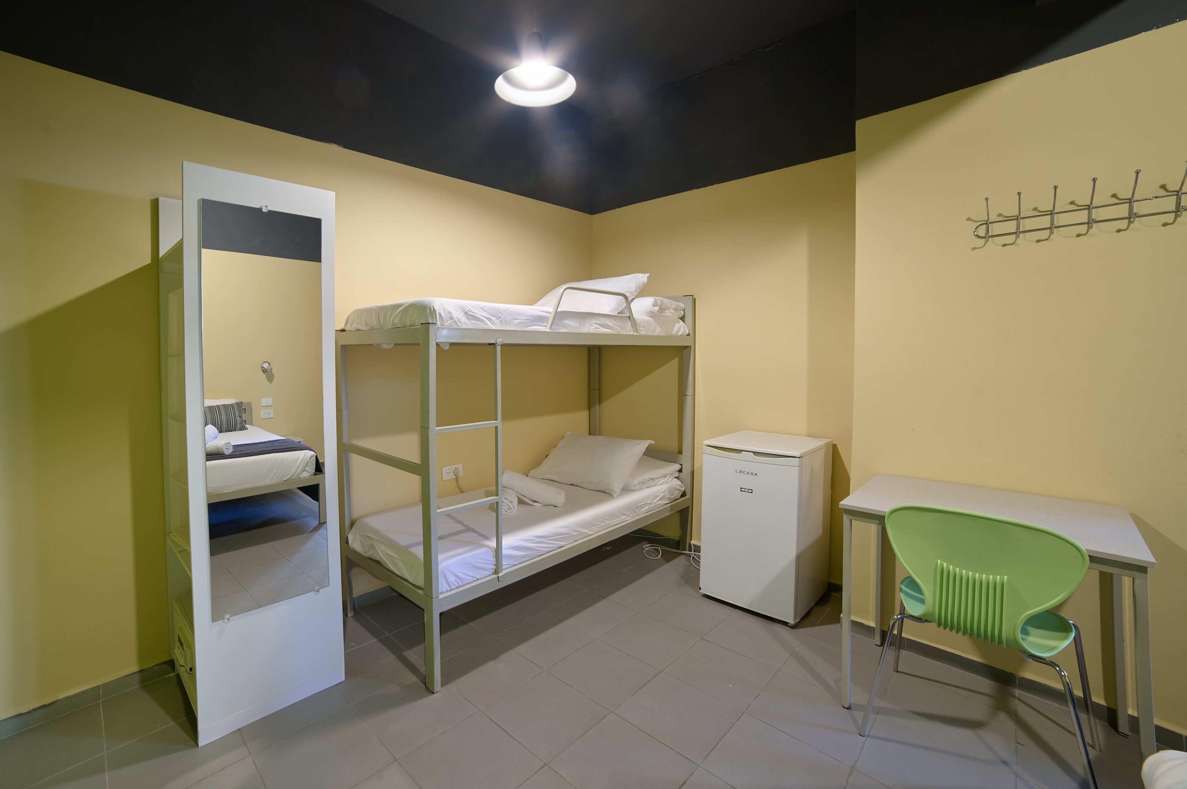A dormitory room with bunk beds, a desk, a chair, and a small refrigerator.