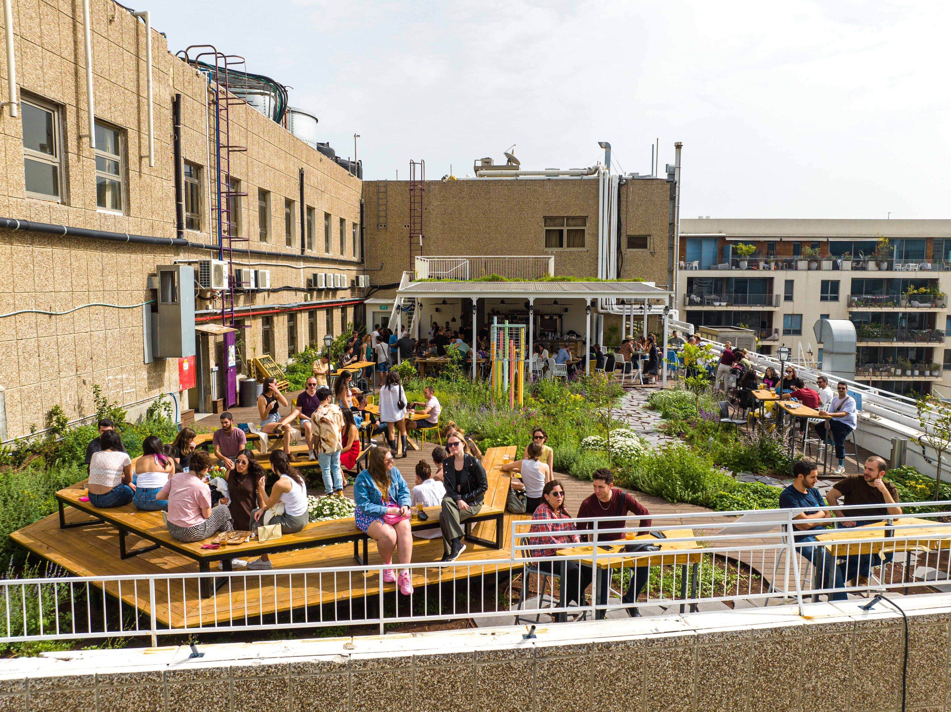 An outdoor dining area with wooden tables and benches, surrounded by greenery and a building with multiple windows and air conditioning units.
