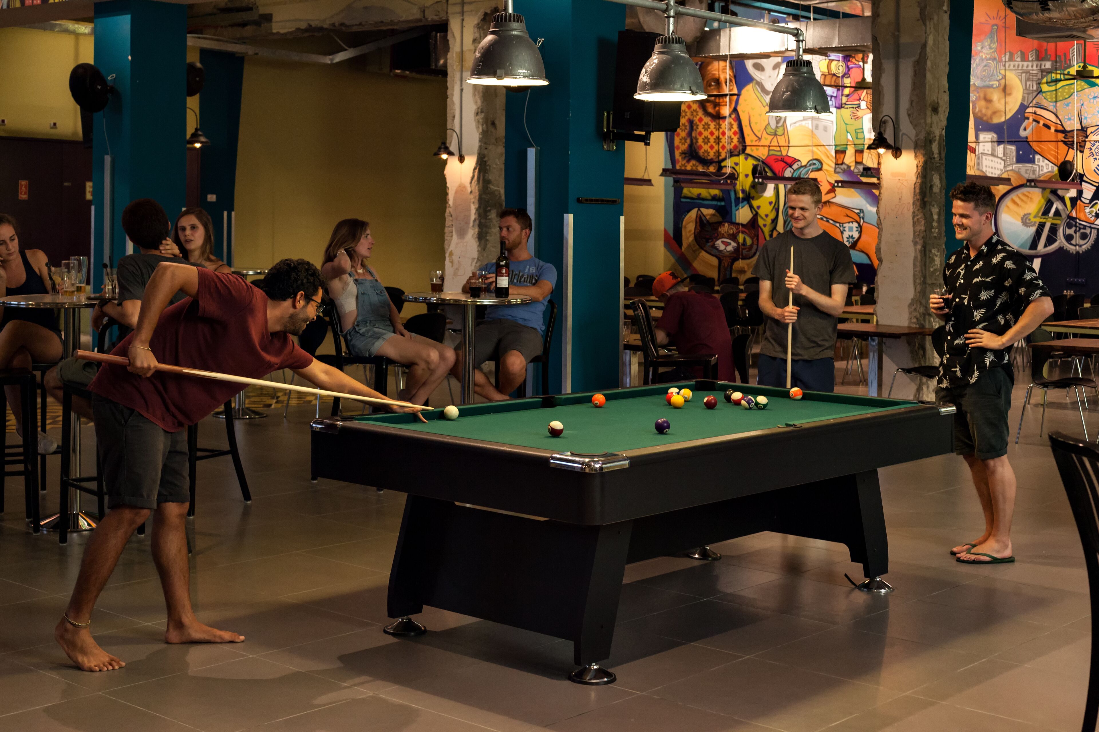 A pool table in a room with a mural on the wall and a group of people around it.