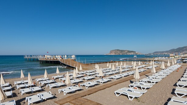 A beach with rows of sun loungers and umbrellas, a pier extending into the sea, and a mountainous backdrop. - Asia Beach Resort & Spa Hotel (Alanya)