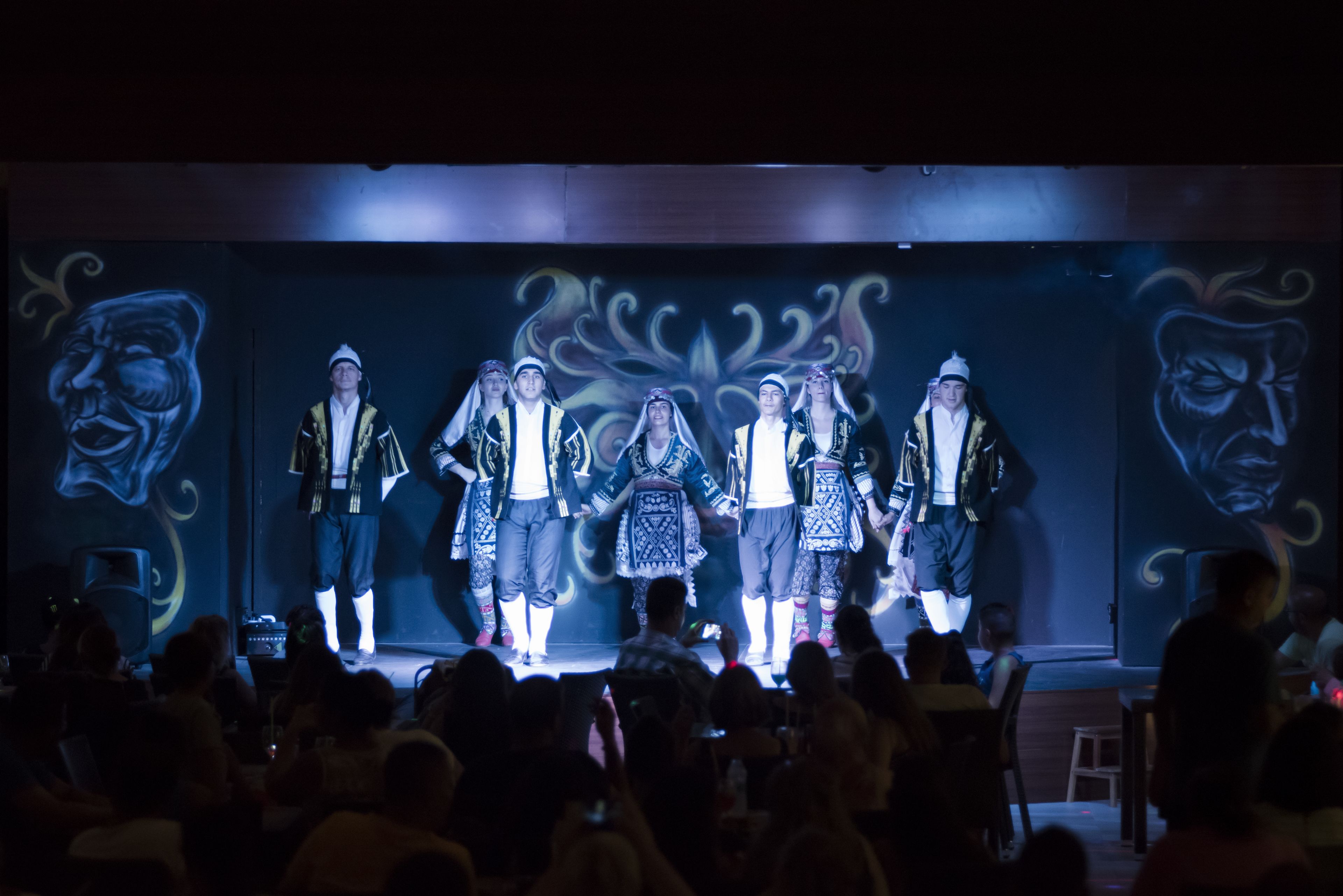A stage with performers in traditional costumes, an audience in the foreground, and a mural on the wall.