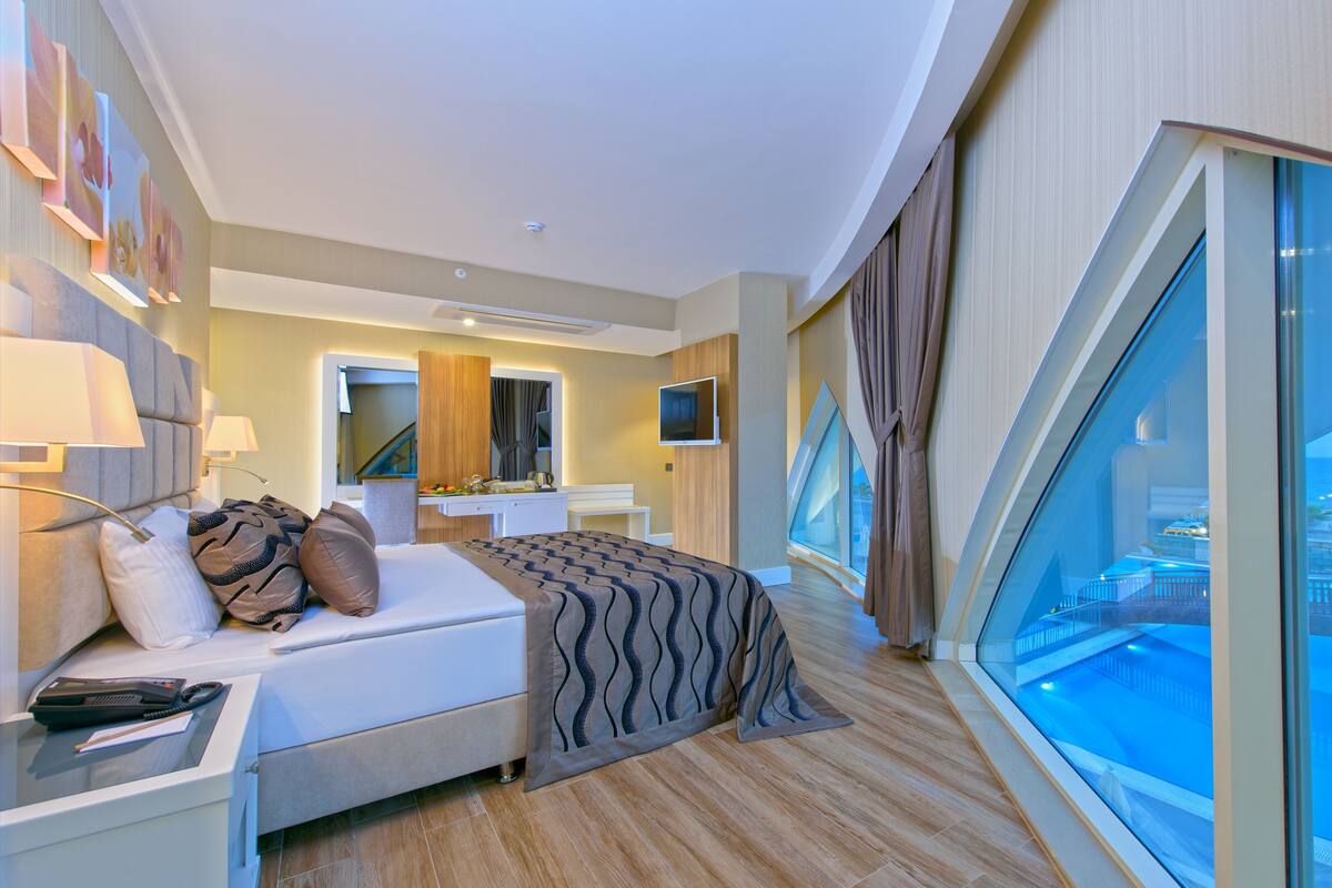 Una habitación de hotel moderna con una cama grande, mesitas de noche, un escritorio, un televisor y un ventanal con vistas a la ciudad.