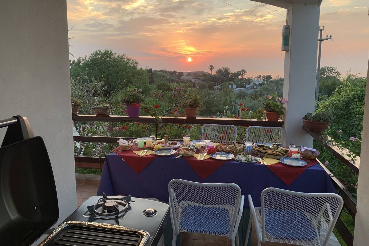 A balcony table set for a meal with a sunset view, surrounded by potted plants and a grill.