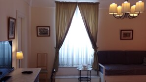 Quarto de hotel com cama, escrivaninha, cadeira, mesa pequena, televisão e janela com cortinas.