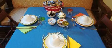 A dining table set with plates, cups, and cutlery, covered with a blue tablecloth, and a basket of folded napkins.