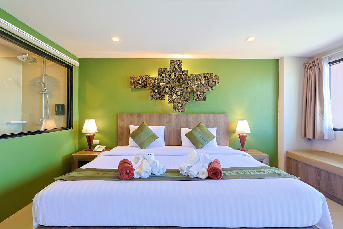 Quarto de hotel com uma cama grande, duas mesas de cabeceira, chuveiro e uma parede verde com padrão decorativo em cruz.