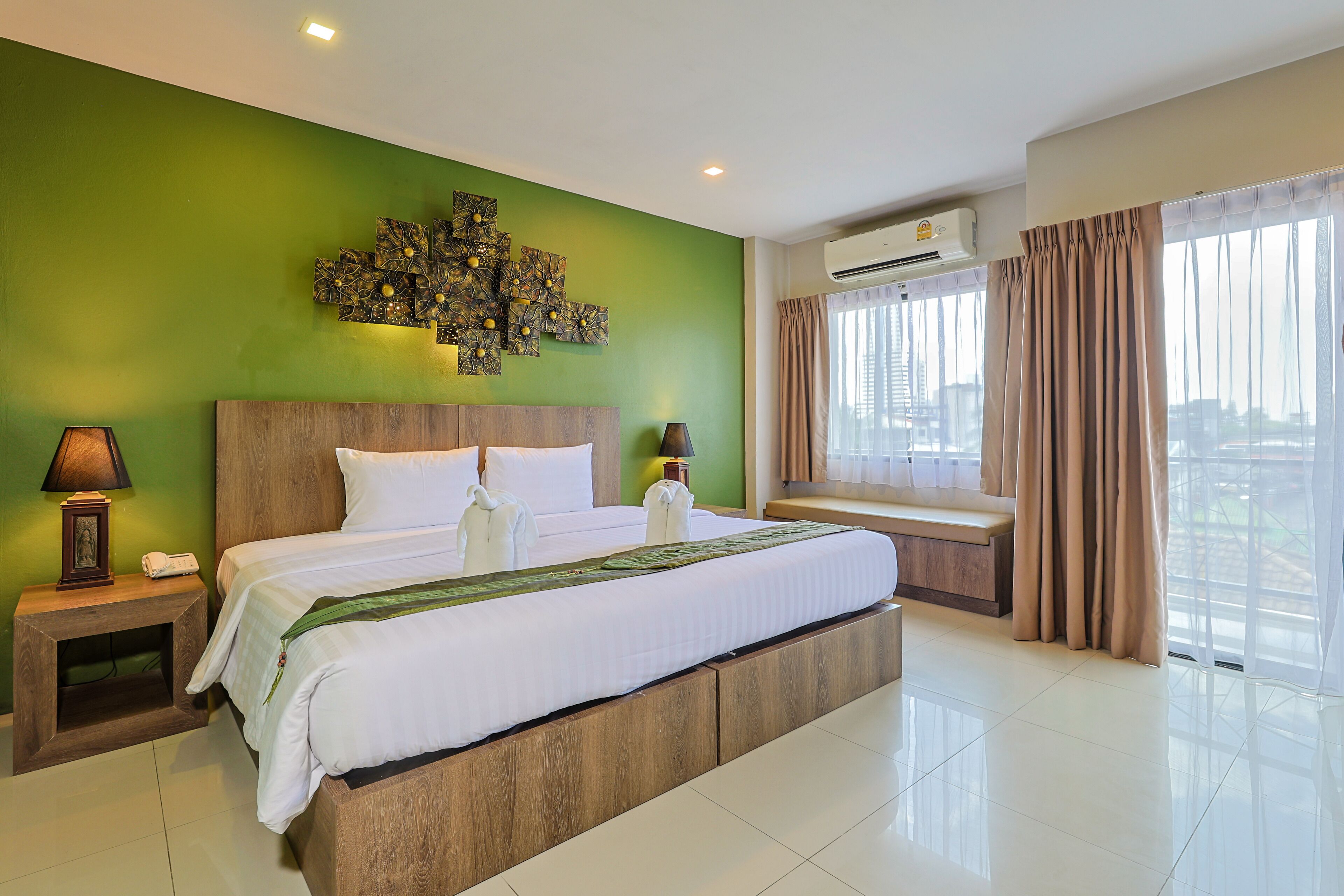 Una habitación de hotel con una cama grande, un cabecero de madera, una pared de acento verde, una ventana con cortinas y un equipo de aire acondicionado.