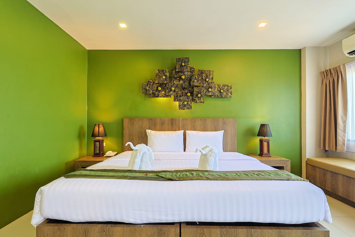 Quarto de hotel com uma cama grande, paredes verdes, cabeceira de madeira, duas luminárias de apoio e um objeto decorativo sobre a cama.