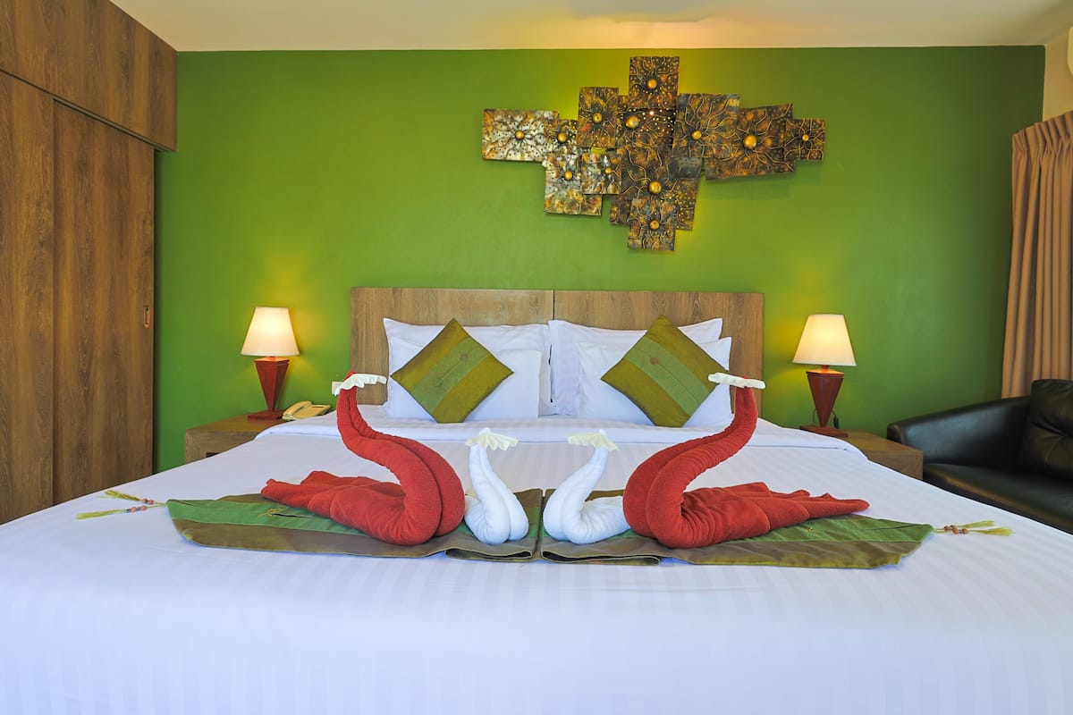 Quarto de hotel com parede verde, cabeceira de madeira, duas luminárias e cama com almofadas em forma de cisne vermelhas e brancas.