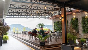 An outdoor patio with tables and chairs, a stone wall with greenery, and a view of mountains. - The Gig Hotel (Patong)