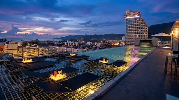 A rooftop pool area with multiple lounge chairs and tables, a large building with a unique shape in the background, and a clear view of the surrounding cityscape. - The Gig Hotel (Patong)