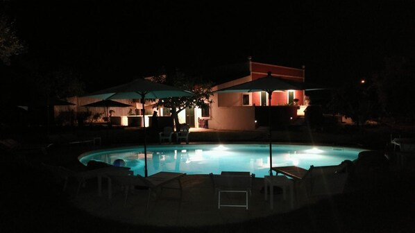 A nighttime view of a swimming pool area with lounge chairs and umbrellas, surrounded by buildings with warm lighting. - B&B Villa Giorgia (Monopoli)